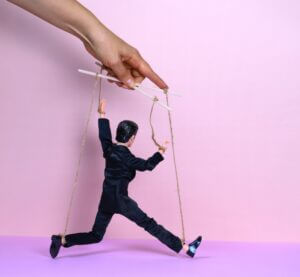 A puppet in a suit controlled by strings on a pink background, symbolizing leadership dynamics, available at Elevate Your Organization's Culture and Performance With CEEK.